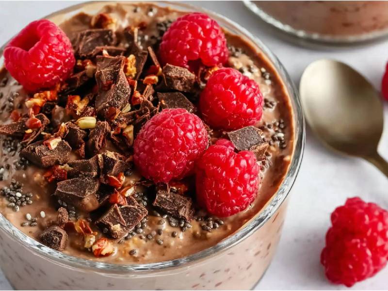 Schale mit Schokoladen-Overnight-Oats mit Himbeeren, Löffel und ein paar frische Himbeeren rechts daneben