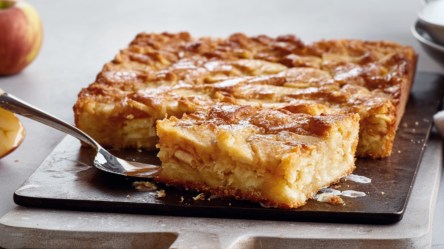 Ein Stück schwedischer Apfelkuchen vom Blech liegt vor dem restlichen Kuchen auf einer Platte.