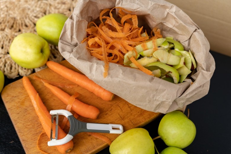 Eine Papiertüte mit Gemüseabfällen, daneben liegen geschälte Karotten und Äpfel. Ein Sparschäler ist ebenfalls zu sehen.