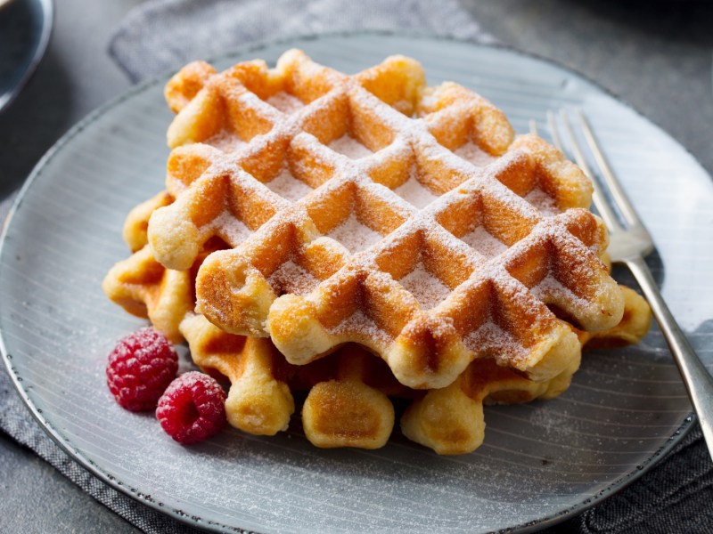 Zwei Skyr-Waffeln übereinander gestapelt auf einem Teller. Daneben liegen frische Himbeeren und sie sind mit Puderzucker bestreut.