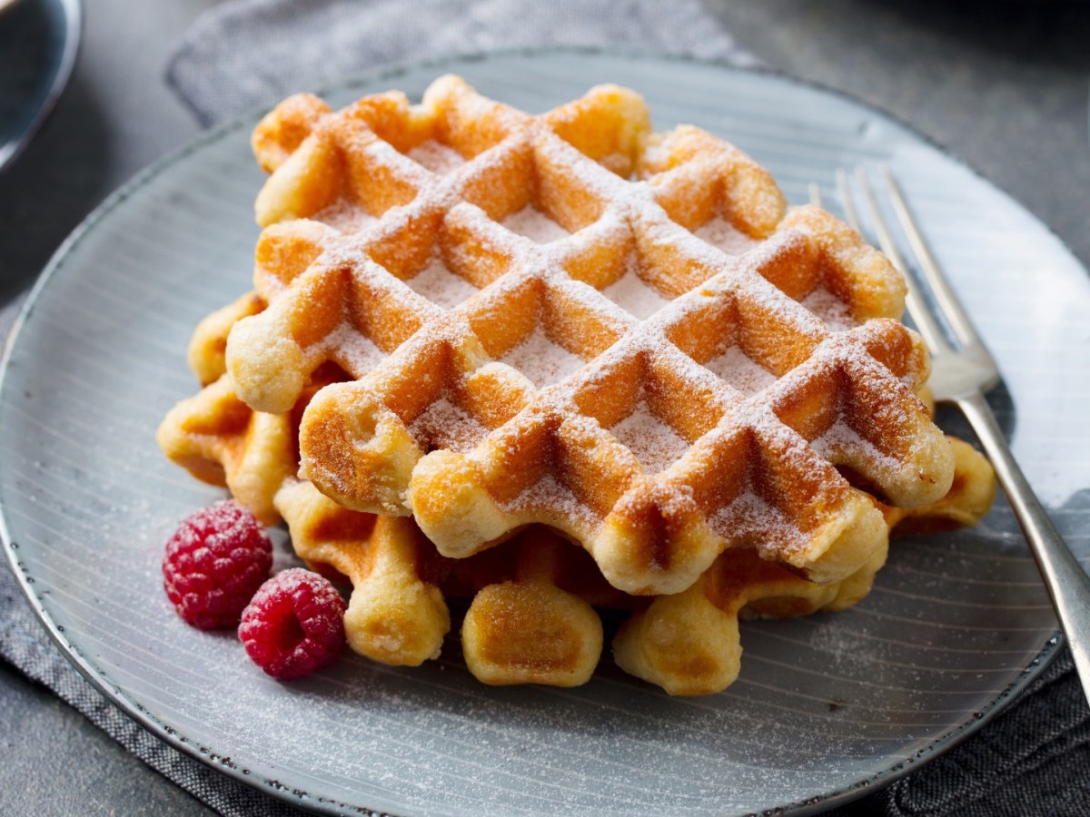 Zwei Skyr-Waffeln übereinander gestapelt auf einem Teller. Daneben liegen frische Himbeeren und sie sind mit Puderzucker bestreut.