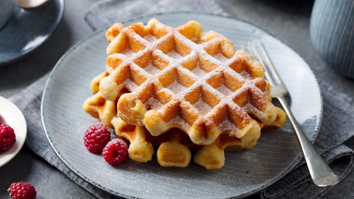 Zwei Skyr-Waffeln übereinander gestapelt auf einem Teller. Daneben liegen frische Himbeeren und sie sind mit Puderzucker bestreut.