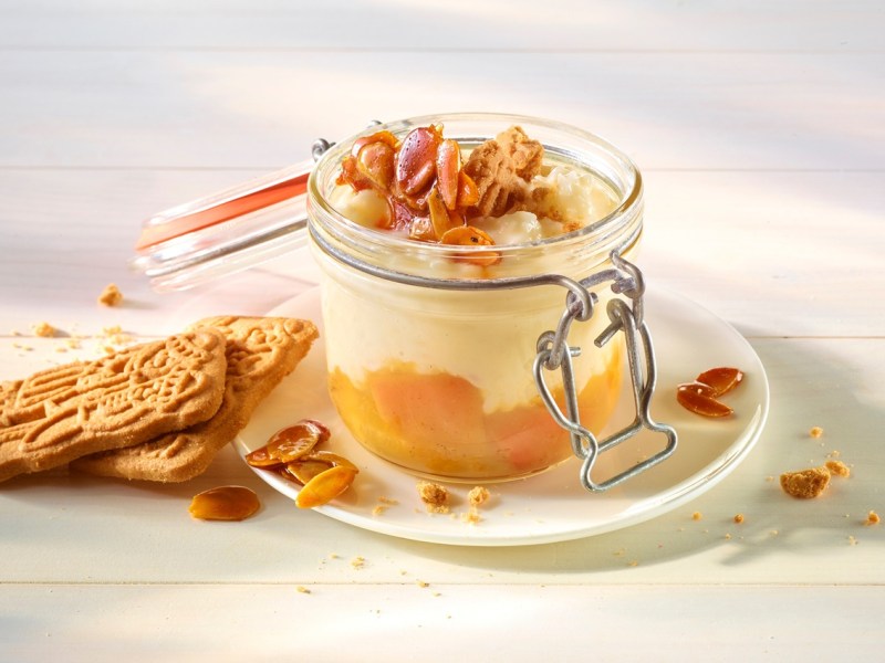 Ein geöffnetes Weckglas gefüllt mit Spekulatius-Milchreis und einer Fruchtschicht. Dekoriert mit Spekulatius-Splittern. Das Glas steht auf einer Untertasse.