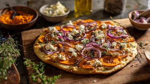 Ein Süßkartoffel-Flammkuchen, der auf einem Holzbrett liegt. Ringsum sind frische Zutaten zu sehen.