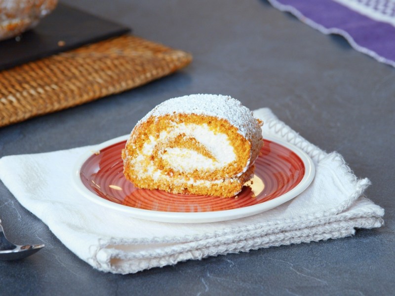 Ein Stück Kürbisrolle, serviert auf einem Teller. Der Teller steht auf einer weißen Stoffserviette, daneben liegt eine Kuchengabel.