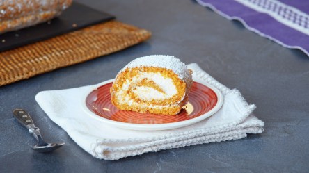 Ein Stück Kürbisrolle, serviert auf einem Teller. Der Teller steht auf einer weißen Stoffserviette, daneben liegt eine Kuchengabel.
