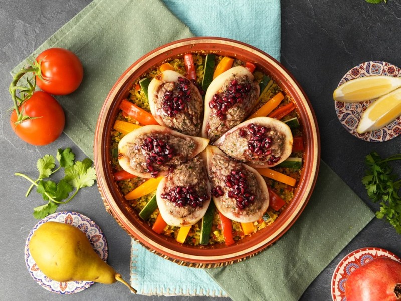 Eine Tajine, gefüllt mit herzhaft gefüllten Birnen. Außen herum liegen Tomaten, Birnen, Zitrone und ein Granatapfel als Deko.