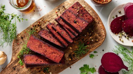 Ein Holzbrett mit einigen Scheiben Tofu-Pastrami, das mit Gewürzen garniert ist. Rechts kann man eine Schale mit aufgeschnittener Rote Bete erkennen. Außerdem liegen frische Kräuter verstreut.