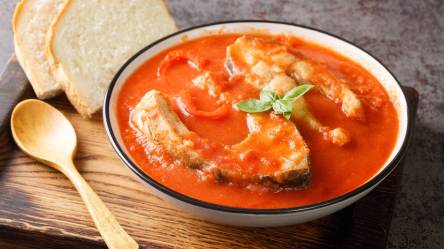 Ein Teller mit ungarischer Fischsuppe. Der Teller steht auf einem Holzbrett und ein Holzlöffel liegt dabei. Die Suppe wird mit zwei Scheiben Weißbrot serviert.