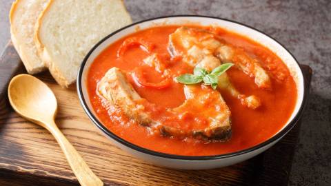 Ein Teller mit ungarischer Fischsuppe. Der Teller steht auf einem Holzbrett und ein Holzlöffel liegt dabei. Die Suppe wird mit zwei Scheiben Weißbrot serviert.