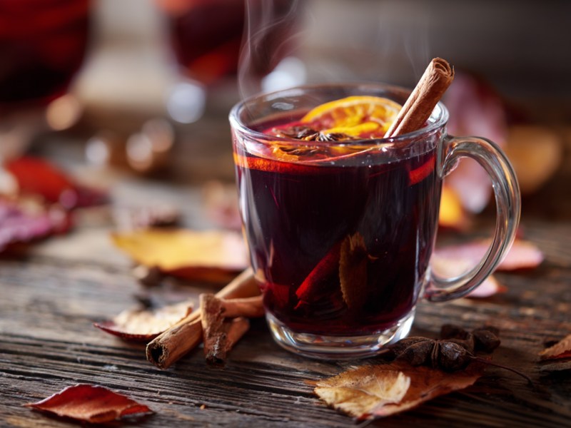 Eine Glastasse mit Zwetschgen-Punsch. Im Punsch schwimmen einige Ingwer- und Orangenscheiben sowie eine Zimtstange als Dekoration. Die Tasse steht auf einem rustikalen Holztisch, drum herum liegen Blätter.