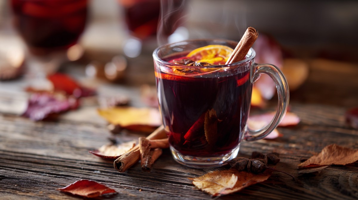 Eine Glastasse mit Zwetschgen-Punsch. Im Punsch schwimmen einige Ingwer- und Orangenscheiben sowie eine Zimtstange als Dekoration. Die Tasse steht auf einem rustikalen Holztisch, drum herum liegen Blätter.