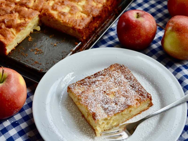 Ein Apfel-Marzipan-Kuchen vom Blech, von dem ein Stück abgeschnitten wurde, welches jetzt auf einem Teller davor liegt.