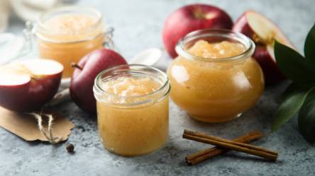 Drei Gläser Apfel-Marzipan-Marmelade auf einem Tisch, Zimtstangen und Äpfel daneben
