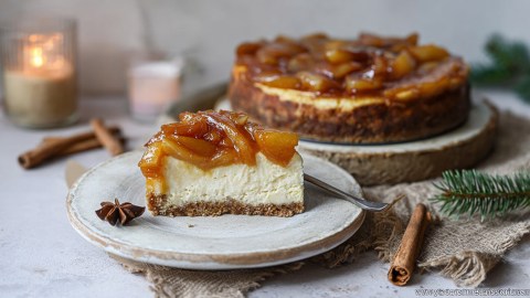 Ein Stück Bratapfel-Cheesecake, das auf einem Kuchenteller liegt. Daneben liegt ein Sternanis. Im Hintergrund kann man einen ganzen Bratapfel-Cheesecake erkennen. Ringsum liegen einige Tannenzweige, Zimtstangen und Kerzen. Weihnachtliches, gemütliches Ambiente.