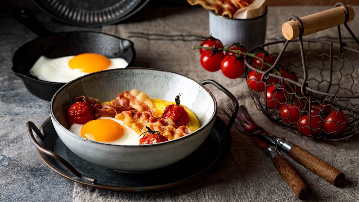 Eine Schale Frühstücks-Polenta mit Ei und Bacon, daneben Kirschtomaten.