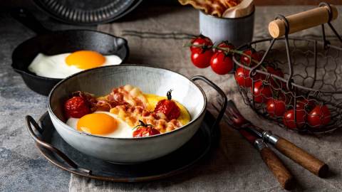 Eine Schale Frühstücks-Polenta mit Ei und Bacon, daneben Kirschtomaten.