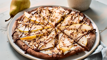 Ein großer Teller mit Frühstückspizza mit Birnen und Mandeln. Im Hintergrund liegen zwei Birnen als Dekoration.