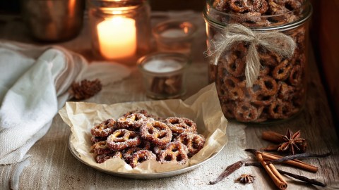 Ein Teller mit Backpapier, auf dem ein kleiner Haufen gebrannte Brezeln liegen. Im Hintergrund ist eine brennende Kerze zu sehen, genauso wie ein großes Glas mit vielen gebrannten Brezeln. Außerdem liegen Gewürze wie Nelken, Vanilleschoten und Zimtstangen herum.