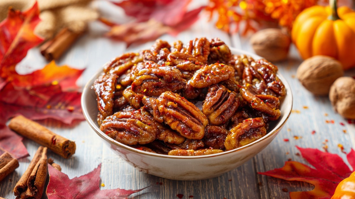 Eine Schüssel voller gerösteter Pekannüsse mit Ahornsirup und Chili. Ringsum liegen Herbstblätter, Zimtstangen und Walnüsse herum.