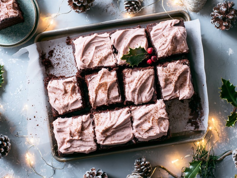 Draufsicht: ein Glühweinkuchen vom Blech mit Frosting. Ringsum liegen Kiefernzapfen und Stechpalmenzweige. Außerdem liegt eine Lichterkette drapiert rund ums Blech. Weihnachtliches Ambiente.