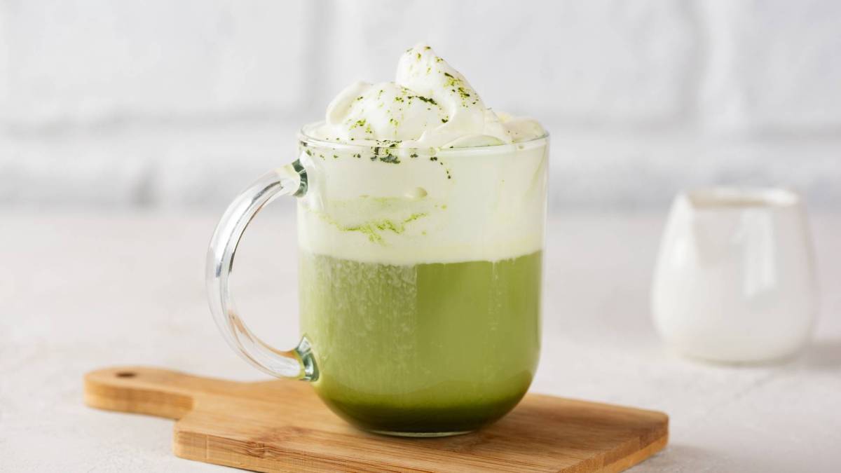 Eine gläserne Tasse heiße weiße Schokolade mit Matcha, die auf einem kleinen Holzbrett steht vor einem hellen, minimalistischen Hintergrund.