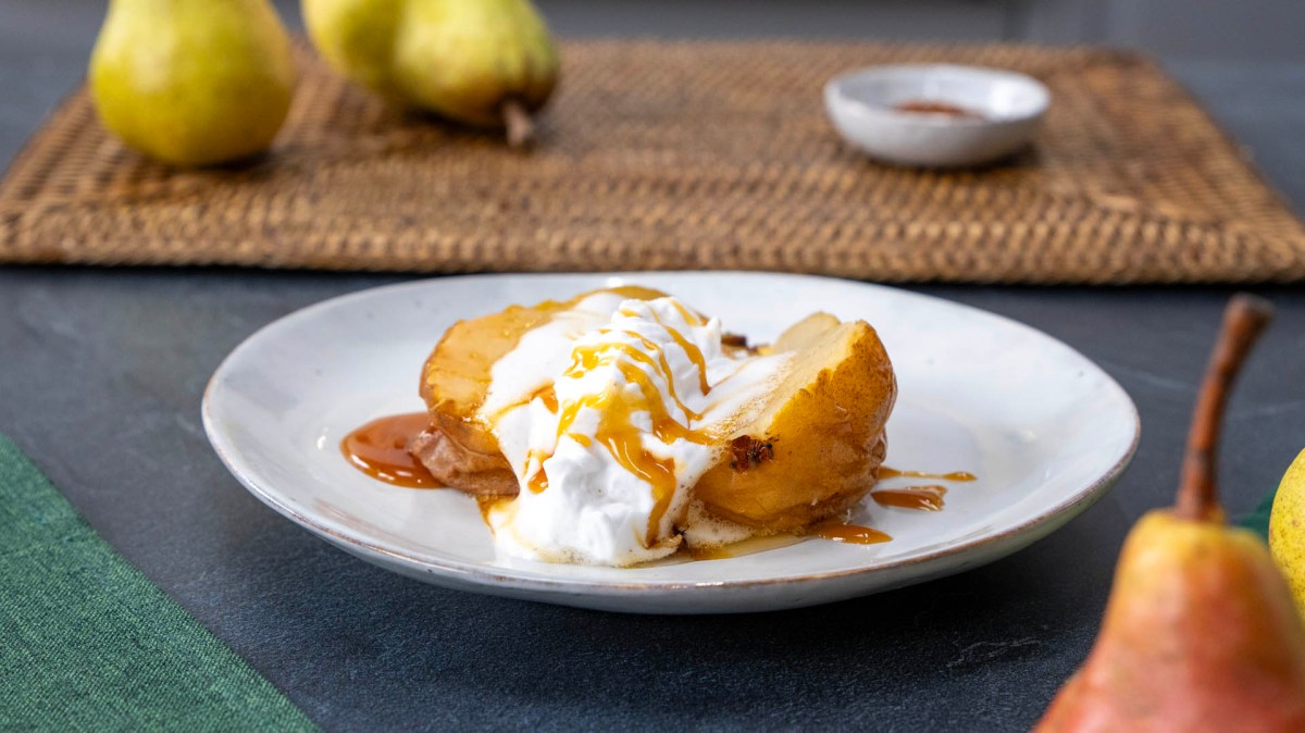 Ein Teller mit einer angerichteten holländischen Birne. Im Hintergrund kann man auf einem Tischuntersetzer Birnen und eine Schale mit Gewürz erkennen. Im Vordergrund ragen auch Birnen ins Bild.