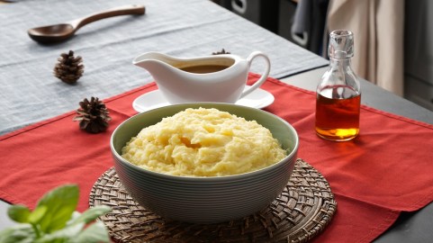 Eine Schüssel mit Kartoffelpüree mit Bratensoße steht auf einem Brett. Ein Tisch ist hübsch gedeckt mit roter Tischdecke und Besteck. Im Hintergrund steht eine Soßenschale.