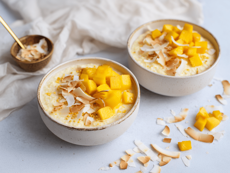 Zwei Schüssel Kokos-Mano-Porridge auf einer grauen Unterlage. Vor ihnen liegen mehrere Kokosraspeln sowie einige Mangowürfel.
