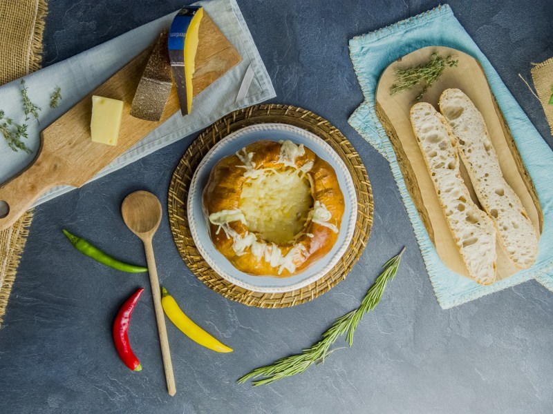 Draufsicht: Kürbis-Käse-Fondue. Daneben liegt ein Brett mit einem aufgeschnittenen Baguette, ein Brett mit Käsestücken sowie ein Holzlöffel und einige Chilischoten verteil.