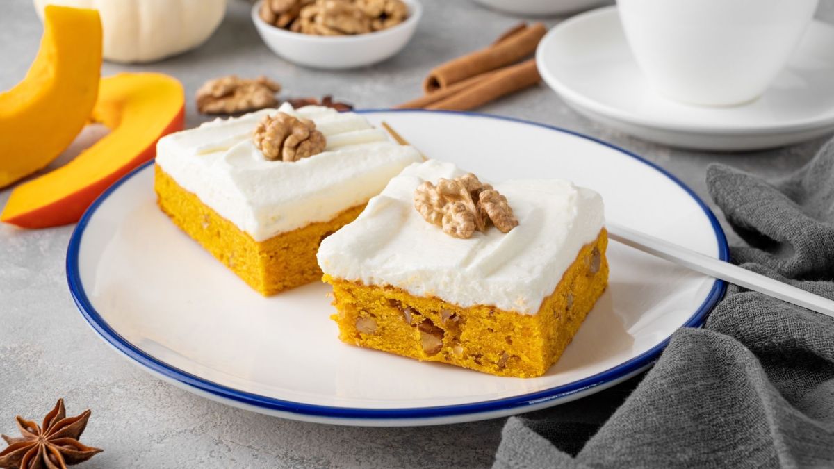 Zwei Stücke Kürbis-Walnuss-Kuchen vom Blech auf einem weißen Teller mit blauem Rand. Daneben liegen Kürbisspalten und Gewürze.