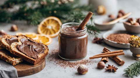 Ein Glas Lebkuchen-Aufstrich mit einer Zimtstange zum Umrühren drin. Neben dem Glas steht ein Brett mit einigen Schreiben Brot, die mit dem Aufstrich bestrichen sind. Ansonsten liegen Zimtstangen, Sternanis und Haselnüsse herum. Auch eine Schale mit Kakaopulver ist zu sehen.