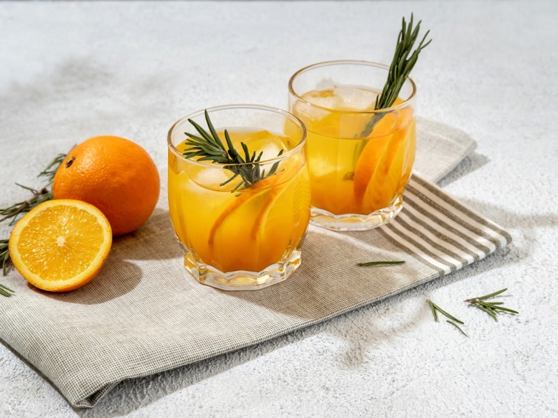 Zwei Gläser mit Mandarinen-Lillet mit Zimt stehen auf einer Stoffserviette. Garniert sind sie mit zwei Rosmarinzweigen. Eine aufgeschnittene Orange und ganze Orange liegen daneben.