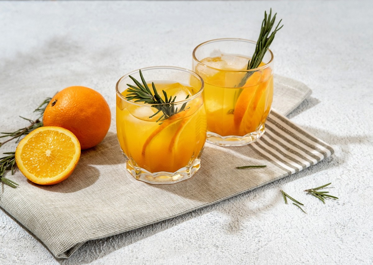 Zwei Gläser mit Mandarinen-Lillet mit Zimt stehen auf einer Stoffserviette. Garniert sind sie mit zwei Rosmarinzweigen. Eine aufgeschnittene Orange und ganze Orange liegen daneben.