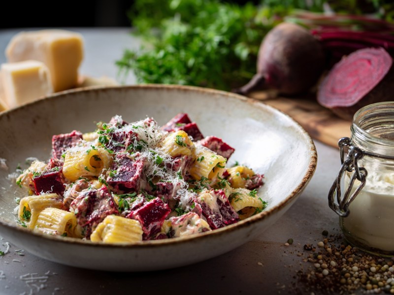 Ein Teller Pasta mit Roter Bete und Crème fraîche. Ringsum und im Hintergrund kann man frische Rote Bete, Stücke Parmesan und ein Glas mit Crème fraîche erkennen.