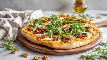 Ein rundes Holzbrett, auf dem eine Pizza mit Kürbissoße, Chorizo und Rucola liegt. Ringsum liegen ein paar Walnüsse und frischer Rucola verstreut.