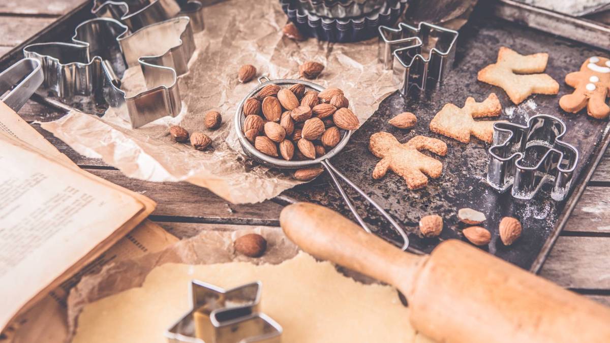 Ein Holztisch, auf dem gerade Plätzchen gebacken werden. Es liegen Teig, Ausstecher, Mehl, Mandeln, Plätzchen, ein Nudelholz, etc. herum.