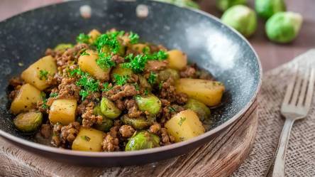 Rosenkohl-Hackfleischpfanne mit Kartoffeln auf einem Tisch, Rosenkohl im Hintergrund und Gabel daneben
