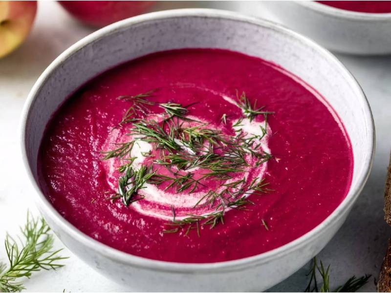 Eine Schale mit Rote-Bete-Cremesuppe mit Apfel auf einem Tisch, Scheibe Brot daneben und Apfel im Hintergrund
