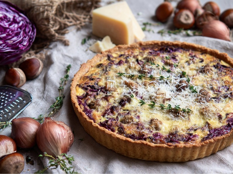 Eine Rotkohl-Maronen-Quiche, die auf einem mit einer Tischdecke bedeckten Tisch liegt. Ringsum kann man einen halben Rotkohl, frische Maronen, ein Stück Parmesan sowie Zwiebeln und Thymianzweige sehen.