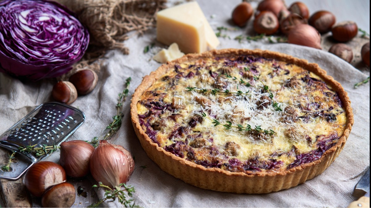 Eine Rotkohl-Maronen-Quiche, die auf einem mit einer Tischdecke bedeckten Tisch liegt. Ringsum kann man einen halben Rotkohl, frische Maronen, ein Stück Parmesan sowie Zwiebeln und Thymianzweige sehen.