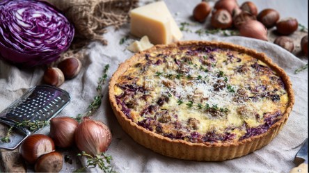 Eine Rotkohl-Maronen-Quiche, die auf einem mit einer Tischdecke bedeckten Tisch liegt. Ringsum kann man einen halben Rotkohl, frische Maronen, ein Stück Parmesan sowie Zwiebeln und Thymianzweige sehen.