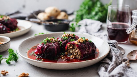 Ein Teller mit Rotkohlrouladen mit Hackfleisch-Nuss-Füllung, die mit etwas Rotweinsoße angerichtet sind. Im Hintergrund kann man Kartoffeln in einer Schale, einen Bund frischer Kräuter, ein Glas mit Rotwein sowie einen weiteren Teller mit Rouladen erkennen.