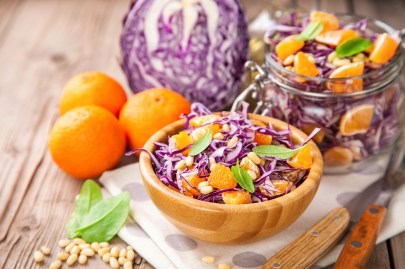 Eine Schüssel voll Rotkohlsalat mit Mandarine. Die Schüssel steht auf einer Stoffserviette. Außen herum liegen Mandarinen und ein Rotkohl. Im Hintergrund steht ein geöffnetetes Bügelglas mit dem Salat.